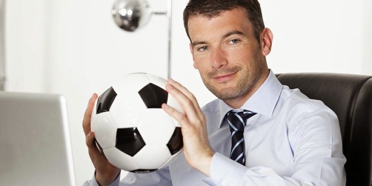 Um homem sentando em seu escritório, de frente para o computador, segurando uma bola de futebol. Como criar um torneio Fantasy