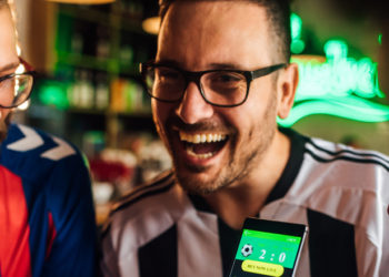 Amigos bebendo cerveja, assistindo jogo de futebol e usando aplicativo móvel para palpites em fantasy