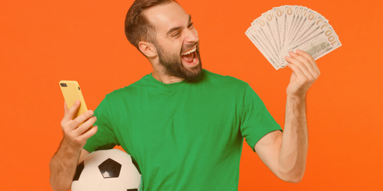 Fã de futebol jovem muito feliz com camiseta verde segurando uma bola de futebol e celular na mão direita e notas de dinehiro na mão esquerda. Ele está em fundo laranja.