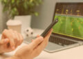 Primeira Aposta: Mão masculina segurando o smartphone. Homem assistindo jogo de futebol, transmissão ao vivo on-line em seu laptop e fazendo apostas no seu time favorito usando aplicativo móvel.